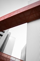 A modern architectural structure featuring tall buildings and a prominent red metal bridge or walkway connecting them. The lines are clean with a focus on industrial design elements, incorporating vertical corrugated panels in a muted gray tone.