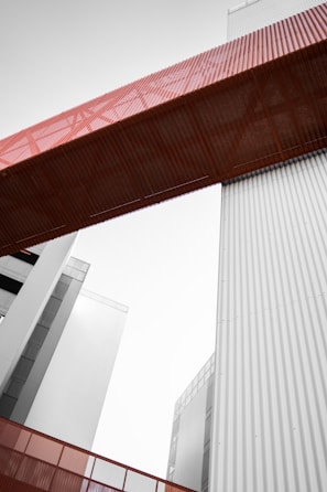 A modern architectural structure featuring tall buildings and a prominent red metal bridge or walkway connecting them. The lines are clean with a focus on industrial design elements, incorporating vertical corrugated panels in a muted gray tone.
