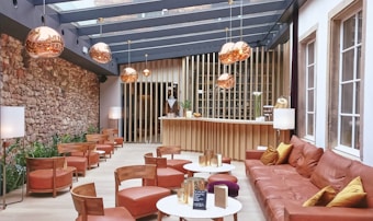 A modern lounge area with plush leather seating and wooden chairs arranged around small white circular tables. The space features a stone wall filled with green plants, and a sleek bar with vertical wooden slats is visible in the background. Large copper pendant lights hang from the ceiling, casting a warm glow. The ceiling is made of glass panels allowing natural light to fill the room.
