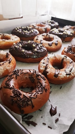 A tray filled with freshly baked donuts covered in chocolate and sprinkles. Some donuts have a glaze, while others are topped with chocolate shavings and decorative sugar balls. They are arranged neatly on parchment paper.