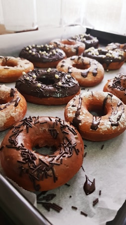 A tray filled with freshly baked donuts covered in chocolate and sprinkles. Some donuts have a glaze, while others are topped with chocolate shavings and decorative sugar balls. They are arranged neatly on parchment paper.