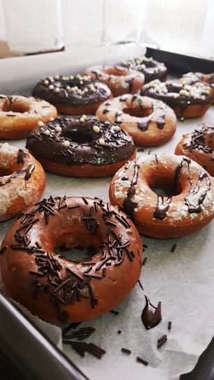A tray filled with freshly baked donuts covered in chocolate and sprinkles. Some donuts have a glaze, while others are topped with chocolate shavings and decorative sugar balls. They are arranged neatly on parchment paper.