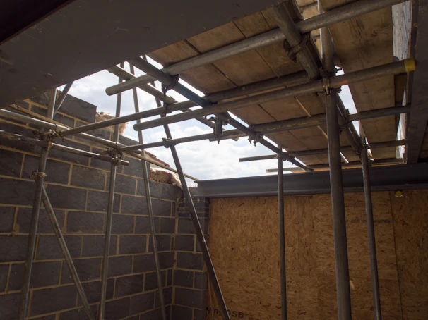 Scaffolding is set up inside a partially constructed building, with metal poles and wooden planks forming platforms. The interior shows exposed brickwork and unfinished walls, with a view of the cloudy sky above through an open section.