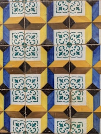 A pattern of decorative ceramic tiles featuring geometric shapes and floral motifs. The design includes repeated squares with alternating blue and yellow triangles, and white squares with green intricate floral patterns. The tiles have a slightly aged and weathered appearance.