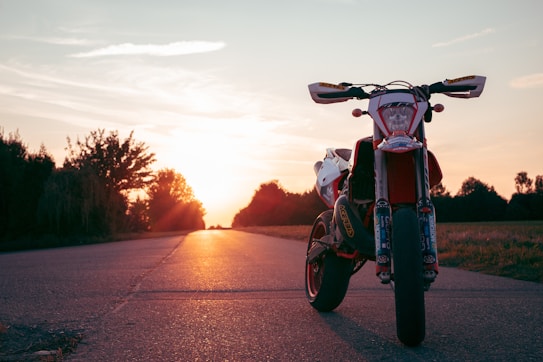 A motorcycle is parked on a deserted road during sunset, casting long shadows. The sky is filled with warm colors as the sun sets on the horizon. The road is flanked by trees on both sides, creating a serene and open atmosphere.