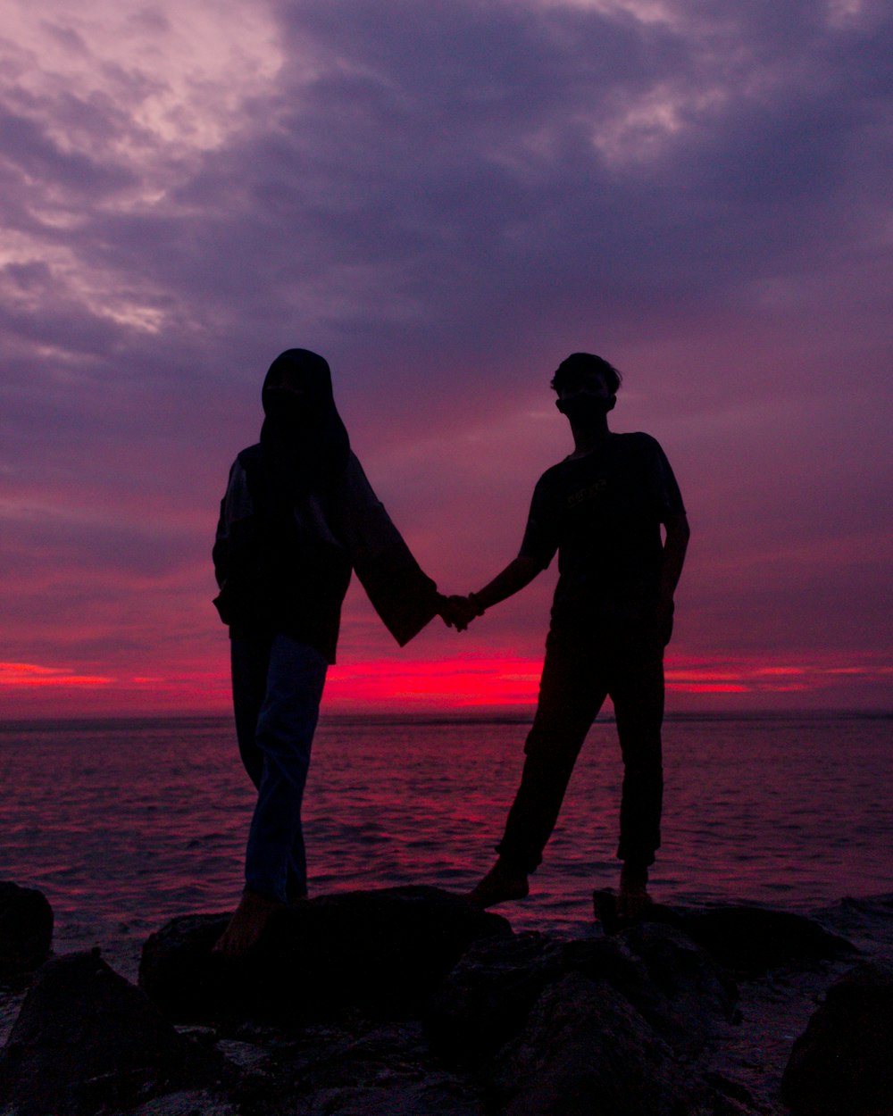 silhouette of man and woman standing on rock near sea during sunset