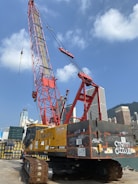 A large red construction crane with a tall boom is situated against a clear blue sky. It is surrounded by high-rise buildings in an urban area. The crane sits on a platform near a waterfront, with graffiti visible on its yellow and black body. Stacks of wooden pallets are nearby.