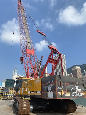 A large red construction crane with a tall boom is situated against a clear blue sky. It is surrounded by high-rise buildings in an urban area. The crane sits on a platform near a waterfront, with graffiti visible on its yellow and black body. Stacks of wooden pallets are nearby.