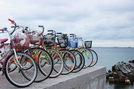 A row of colorful bicycles with baskets lined up on a concrete pier overlooking a calm body of water. The sky is overcast with clouds, casting a serene and subdued atmosphere. There is a collection of rocks to the right, enhancing the coastal setting.