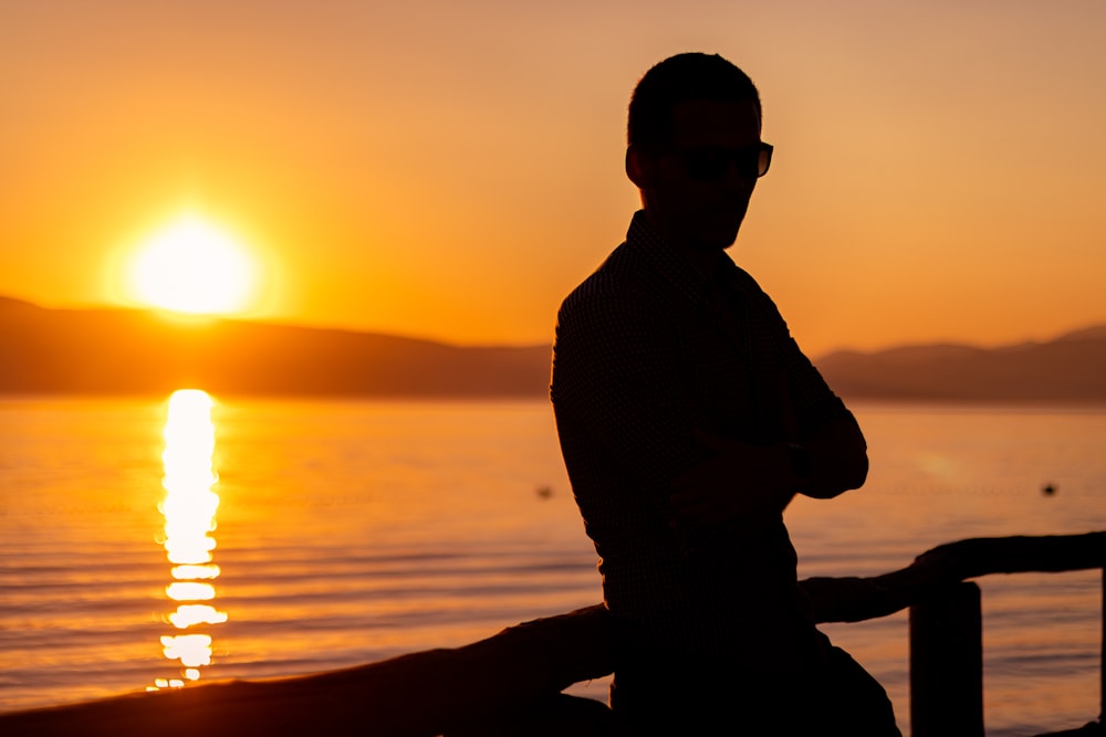 silhouette of man wearing sunglasses and cap sitting on beach during sunset