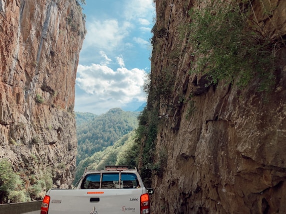 A white pickup truck travels through a narrow mountain pass, flanked by steep, rugged cliffs on both sides. The sky above is partly cloudy, providing a glimpse of lush green forested hills in the distance.