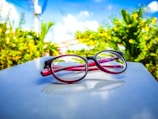 Red and black eyeglasses rest on a white surface, set against a vibrant outdoor background with lush green foliage and a clear blue sky with white clouds.
