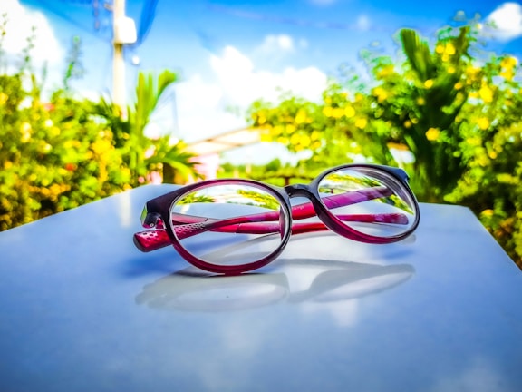 Red and black eyeglasses rest on a white surface, set against a vibrant outdoor background with lush green foliage and a clear blue sky with white clouds.