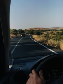A hand is on the steering wheel of a car driving on a rural road. The road stretches ahead, winding slightly with two lanes and white dividing lines. Vegetation, including shrubs and sparse trees, lines the sides of the road. The landscape features rolling hills and a clear sky, giving an open, expansive view.