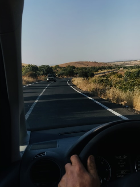 A hand is on the steering wheel of a car driving on a rural road. The road stretches ahead, winding slightly with two lanes and white dividing lines. Vegetation, including shrubs and sparse trees, lines the sides of the road. The landscape features rolling hills and a clear sky, giving an open, expansive view.