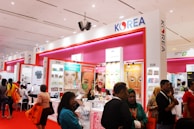 A busy exhibition booth showcasing various Korean beauty products. The booth has bright signage with posters advertising facial treatment creams and masks. People are gathered around, engaging in conversations and exploring the displays. The vibrant red and pink colors are prominent in the booth decor.