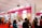 A busy exhibition booth showcasing various Korean beauty products. The booth has bright signage with posters advertising facial treatment creams and masks. People are gathered around, engaging in conversations and exploring the displays. The vibrant red and pink colors are prominent in the booth decor.