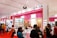 A busy exhibition booth showcasing various Korean beauty products. The booth has bright signage with posters advertising facial treatment creams and masks. People are gathered around, engaging in conversations and exploring the displays. The vibrant red and pink colors are prominent in the booth decor.