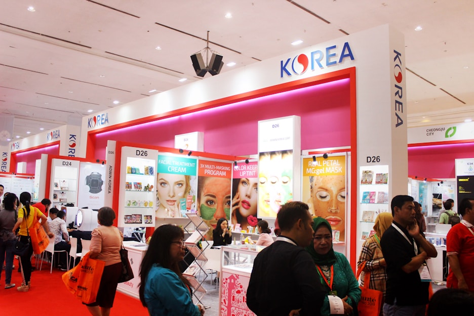 A busy exhibition booth showcasing various Korean beauty products. The booth has bright signage with posters advertising facial treatment creams and masks. People are gathered around, engaging in conversations and exploring the displays. The vibrant red and pink colors are prominent in the booth decor.