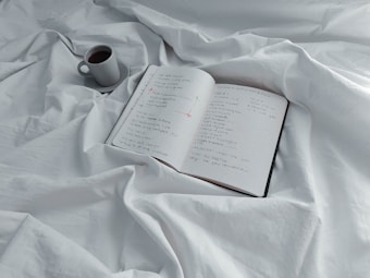 An open notebook with handwritten notes sits on a white, crumpled bedsheet. Next to the notebook is a small mug filled with coffee.
