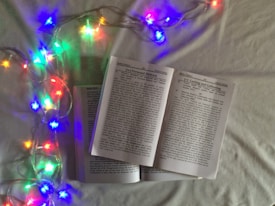 An open book lies on a light-colored surface, surrounded by colorful string lights that emit a warm ambiance. The lights are shaped like stars, glowing in hues of blue, red, green, and yellow, casting gentle reflections on the pages of the book.