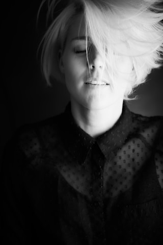 A woman with short blonde hair and closed eyes is captured in a monochrome image. Her hair is tousled, partially covering her face. She wears a dark, semi-transparent shirt with a subtle pattern, creating a contrast against her fair hair and skin.