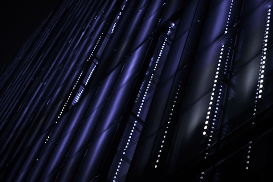 Vertical patterns of blue and purple neon lights glow against a dark background, forming a grid-like structure. The diagonal lines create a sense of depth and modernity.