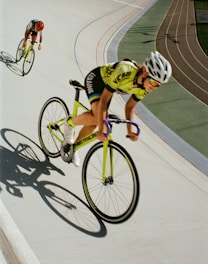 Two cyclists are racing on a velodrome track. The cyclist in the foreground is wearing a yellow and black outfit with a white helmet and is riding a bright yellow bike with purple handlebars. Another cyclist follows closely behind, wearing an orange helmet and black and red outfit, also riding a yellow bike. The velodrome's track is marked with different colored lines, and their shadows are cast on the track.