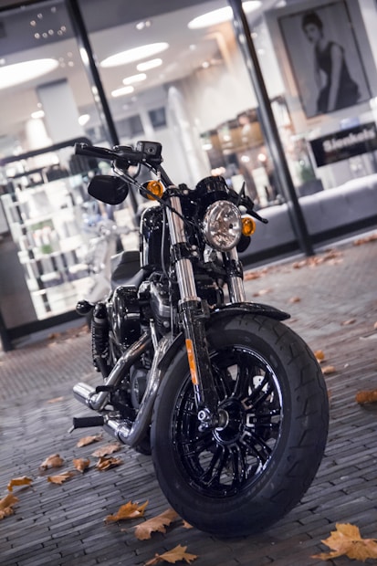 A sleek motorcycle is parked on a cobblestone street, surrounded by fallen autumn leaves. The background features a brightly lit store window displaying various products and a portrait photograph.