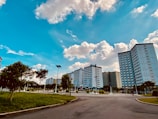 A clean, open urban landscape is seen with tall apartment buildings under a bright blue sky. Fluffy white clouds are scattered across the sky. The foreground shows a paved road and well-maintained green grass areas with trees and lamp posts.