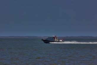 A motorboat speeds across a vast body of water, leaving a white trail behind. The boat has a small cabin and a visible flag at the back. The horizon is flat with distant land, and the sky is clear with a deep blue color.