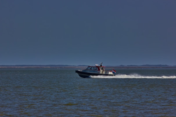 A motorboat speeds across a vast body of water, leaving a white trail behind. The boat has a small cabin and a visible flag at the back. The horizon is flat with distant land, and the sky is clear with a deep blue color.