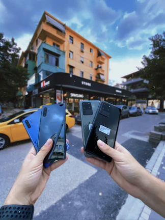 Two hands hold a collection of smartphones in an outdoor urban setting. In the background, a multi-story, colorful building stands near a store with visible signs. Cars, including a yellow one, are parked along the street, trees line the road, and the sky is partly cloudy.