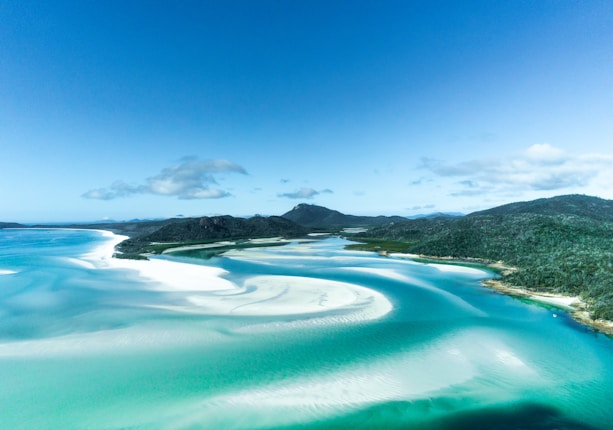 A breathtaking aerial view of a beautiful coastal landscape featuring turquoise waters, pristine white sandy beaches, and lush green hills in the background. The image captures the serene and unspoiled nature of the area, with swirling patterns of sand and water creating a tranquil and picturesque scene under a clear blue sky.