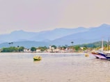 A coastal landscape with calm waters in the foreground and several small boats floating. In the distance, a charming town with white colonial-style buildings and a prominent church is surrounded by lush greenery. Beyond the town, there is a backdrop of majestic, hazy mountains under an overcast sky.