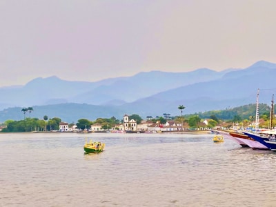 A coastal landscape with calm waters in the foreground and several small boats floating. In the distance, a charming town with white colonial-style buildings and a prominent church is surrounded by lush greenery. Beyond the town, there is a backdrop of majestic, hazy mountains under an overcast sky.