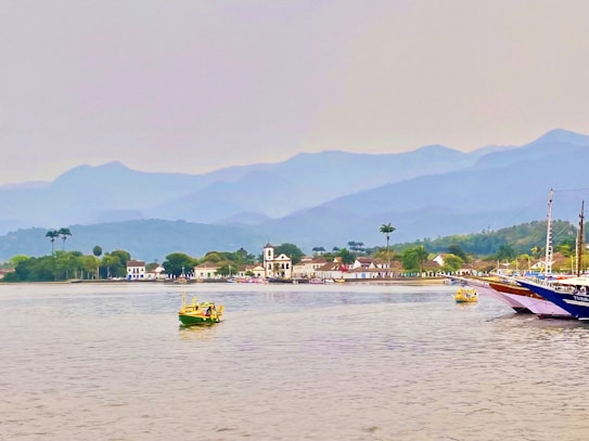 A coastal landscape with calm waters in the foreground and several small boats floating. In the distance, a charming town with white colonial-style buildings and a prominent church is surrounded by lush greenery. Beyond the town, there is a backdrop of majestic, hazy mountains under an overcast sky.