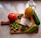 A wooden cutting board sits on a marble countertop. On the board, there is a whole ripe tomato, a green bell pepper, a large cucumber, a few leaves of basil, a cluster of green olives, two slices of white cheese, and a glass bottle with a handle filled with yellow liquid, possibly olive oil.