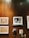 A wall adorned with various artworks and decorations, including a framed black-and-white portrait of two people, textile art pieces with intricate designs, and two masks with a traditional appearance. The wall is wooden, providing a warm, rustic backdrop to the diverse collection.