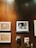 A wall adorned with various artworks and decorations, including a framed black-and-white portrait of two people, textile art pieces with intricate designs, and two masks with a traditional appearance. The wall is wooden, providing a warm, rustic backdrop to the diverse collection.