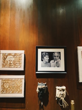 A wall adorned with various artworks and decorations, including a framed black-and-white portrait of two people, textile art pieces with intricate designs, and two masks with a traditional appearance. The wall is wooden, providing a warm, rustic backdrop to the diverse collection.