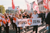 A group of people is participating in a public protest or demonstration, holding a large banner. Several individuals are carrying flags with red and white colors, and there are buildings in the background. Some participants are wearing masks.