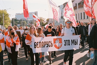 A group of people is participating in a public protest or demonstration, holding a large banner. Several individuals are carrying flags with red and white colors, and there are buildings in the background. Some participants are wearing masks.