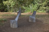 Wooden stumps arranged as outdoor furniture in a natural setting. The seating appears to be crafted from tree trunks placed on a patch of dirt surrounded by grass and foliage. Tall trees and various plants are visible in the background, indicating a forested area.