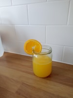 A glass jar filled with orange juice sits on a wooden countertop, with a slice of orange placed on the rim. The background features a white tiled wall.