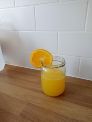 A glass jar filled with orange juice sits on a wooden countertop, with a slice of orange placed on the rim. The background features a white tiled wall.