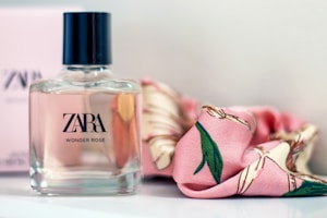 A bottle of Zara Wonder Rose perfume is placed on a surface beside a pink floral-patterned scarf. The bottle has a square shape with a black cap and clear glass revealing a pale pink liquid inside.