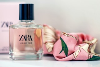 A bottle of Zara Wonder Rose perfume is placed on a surface beside a pink floral-patterned scarf. The bottle has a square shape with a black cap and clear glass revealing a pale pink liquid inside.