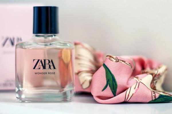A bottle of Zara Wonder Rose perfume is placed on a surface beside a pink floral-patterned scarf. The bottle has a square shape with a black cap and clear glass revealing a pale pink liquid inside.