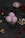 A glass bowl filled with three scoops of pink ice cream, garnished with nuts and rose petals, sits on a dark surface. Scattered cashew nuts and rose petals surround the bowl, with a red rose and another small bowl containing a single scoop of ice cream in the background.